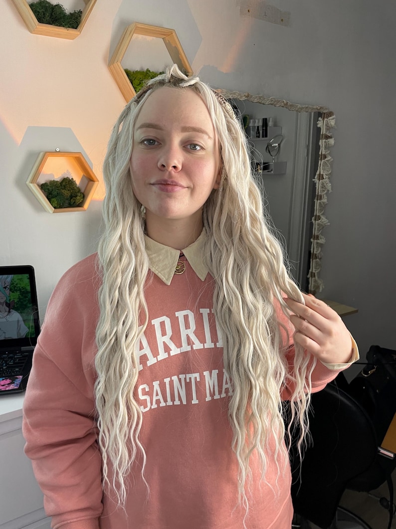 May include: A woman with long, wavy, blonde hair is wearing a pink sweatshirt with the text "Warriors Saint Mary" printed on the front.