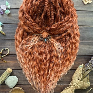 Puede incluir: Una cabeza de rastas de color marrón rojizo con un efecto ombre rubio claro. Las rastas están peinadas con una horquilla decorativa de bronce. El cabello está dispuesto sobre una superficie de madera oscura.