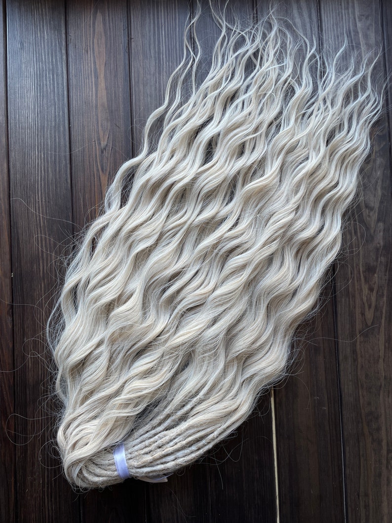 May include: A bundle of long, wavy, white synthetic dreadlocks. The dreadlocks are tied together with a purple ribbon.