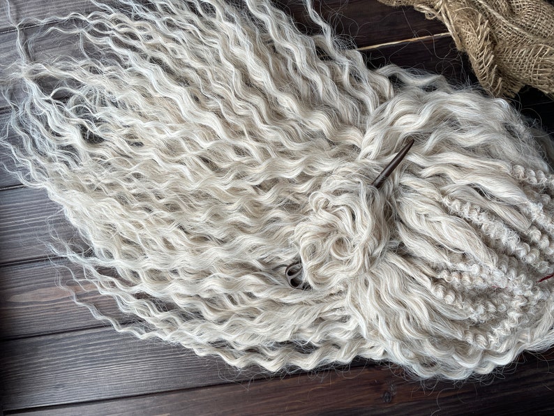 May include: A close-up of a bundle of white curly hair extensions. The hair is arranged in a loose braid and is lying on a dark wooden surface.
