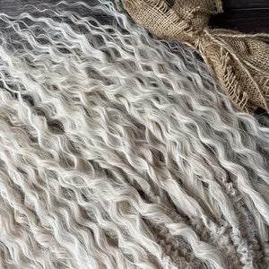 May include: A close-up of a bundle of wavy, light blonde human hair extensions. The hair is soft and textured, with a natural, slightly grayed look.