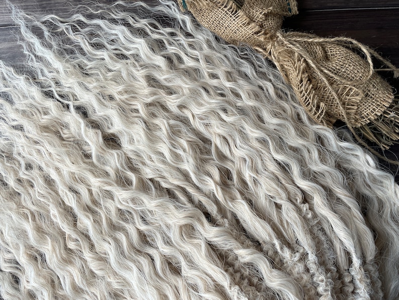 May include: A close-up of a bundle of wavy, light blonde human hair extensions. The hair is soft and textured, with a natural, slightly grayed look.