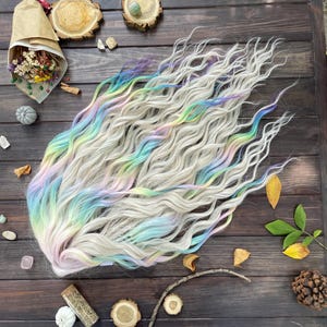 Puede incluir: Un conjunto de extensiones de cabello sintético ondulado gris claro con reflejos de arcoíris pastel. Las extensiones tienen mechones azules, morados, verdes y rosas. El cabello está peinado para que parezca voluminoso y fluido, adecuado para cosplay o moda.