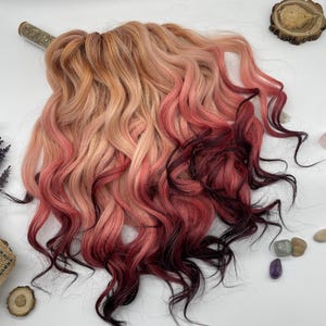 May include: Ombre hair extensions with a blend of colors, transitioning from light peach to pink and deep burgundy. The wavy extensions are styled to showcase the color gradient. The hair is arranged on a white surface with decorative elements.