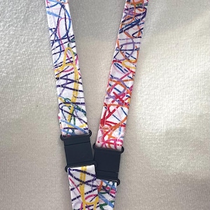 May include: A fabric lanyard featuring a white background with a colorful, abstract scribble pattern in red, blue, yellow, and green. The lanyard includes black plastic side-release buckles and a silver-toned metal swivel lobster claw clasp.
