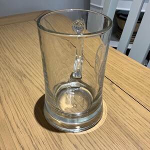 May include: A clear glass beer mug with a handle. The mug has a cylindrical shape and a thick base. The handle is ornate with a decorative design. The mug is sitting on a wooden table.