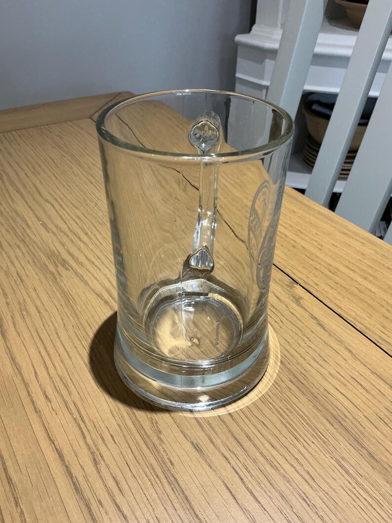 May include: A clear glass beer mug with a handle. The mug has a cylindrical shape and a thick base. The handle is ornate with a decorative design. The mug is sitting on a wooden table.