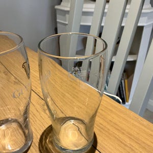 Puede incluir: Dos vasos de pinta transparentes sobre una mesa de madera. Uno tiene grabada la palabra "Guinness". El otro tiene grabada la palabra "PINT" y las letras "M19". Los vasos est&aacute;n vac&iacute;os y proyectan sombras.