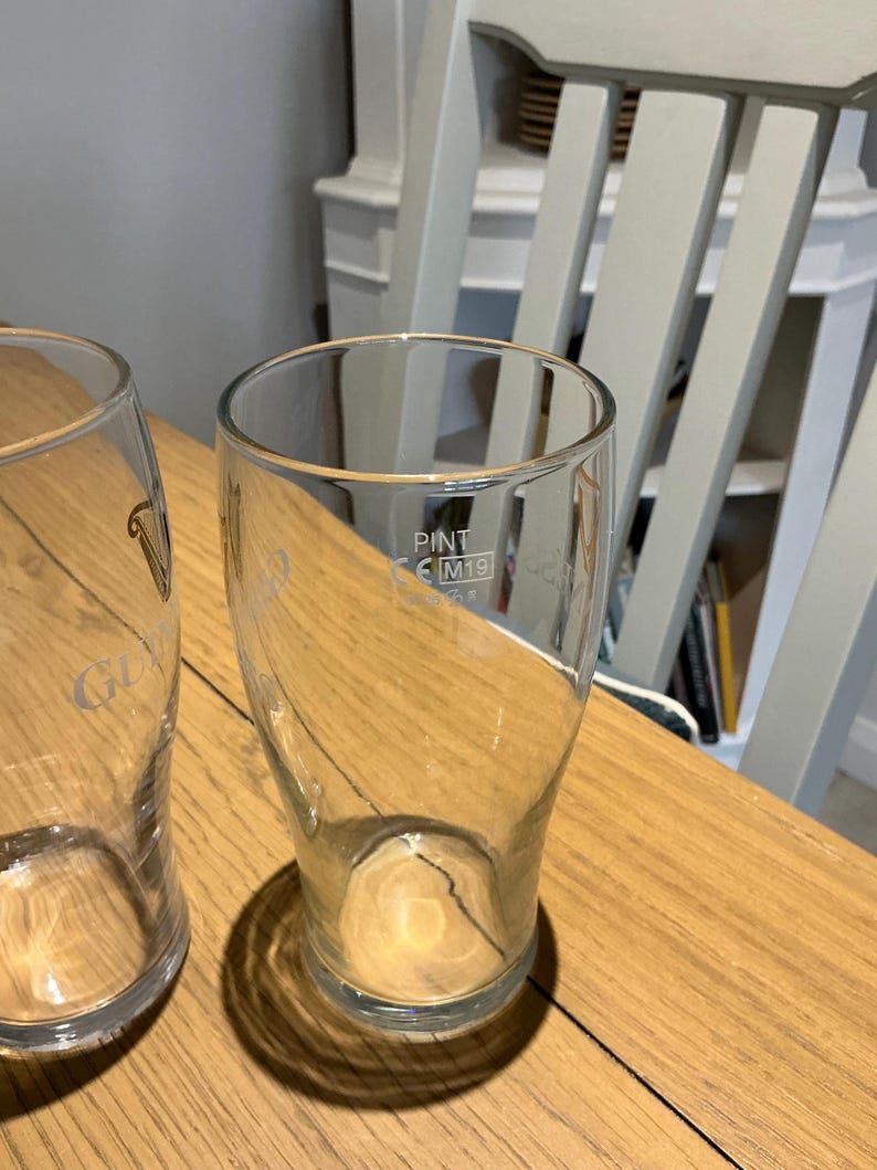 Puede incluir: Dos vasos de pinta transparentes sobre una mesa de madera. Uno tiene grabada la palabra "Guinness". El otro tiene grabada la palabra "PINT" y las letras "M19". Los vasos est&aacute;n vac&iacute;os y proyectan sombras.