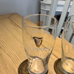 Puede incluir: Vaso de pinta transparente con el logotipo de Guinness en dorado. El vaso tiene una forma curvada y una boca ancha. El logotipo de Guinness presenta un dise&ntilde;o de arpa y la palabra "Guinness". El vaso est&aacute; sobre una mesa de madera.