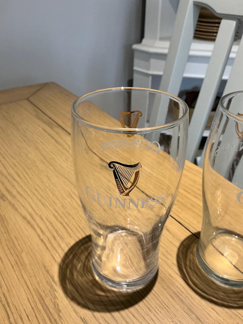 Puede incluir: Vaso de pinta transparente con el logotipo de Guinness en dorado. El vaso tiene una forma curvada y una boca ancha. El logotipo de Guinness presenta un dise&ntilde;o de arpa y la palabra "Guinness". El vaso est&aacute; sobre una mesa de madera.