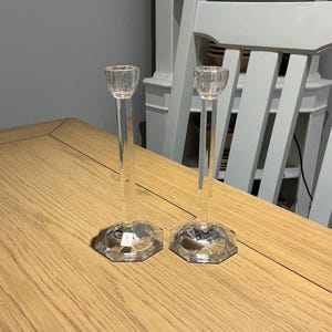 Puede incluir: Dos candelabros de vidrio transparente con bases hexagonales y tallos largos y delgados. Los candelabros están sobre una mesa de madera clara. El fondo incluye una silla blanca y un aparador con platos.