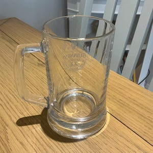 May include: A clear glass beer mug with a handle. The mug has a cylindrical shape and a thick base. The Guinness logo is visible on the front of the mug. The mug is sitting on a wooden table.