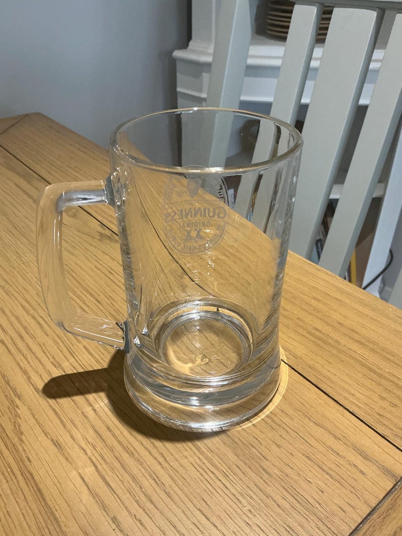 May include: A clear glass beer mug with a handle. The mug has a cylindrical shape and a thick base. The Guinness logo is visible on the front of the mug. The mug is sitting on a wooden table.