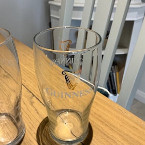 Puede incluir: Vaso de pinta transparente con el logotipo de Guinness, con un arpa dorada y la palabra "Guinness". El vaso est&aacute; sobre una superficie de madera, con un segundo vaso parcialmente visible.