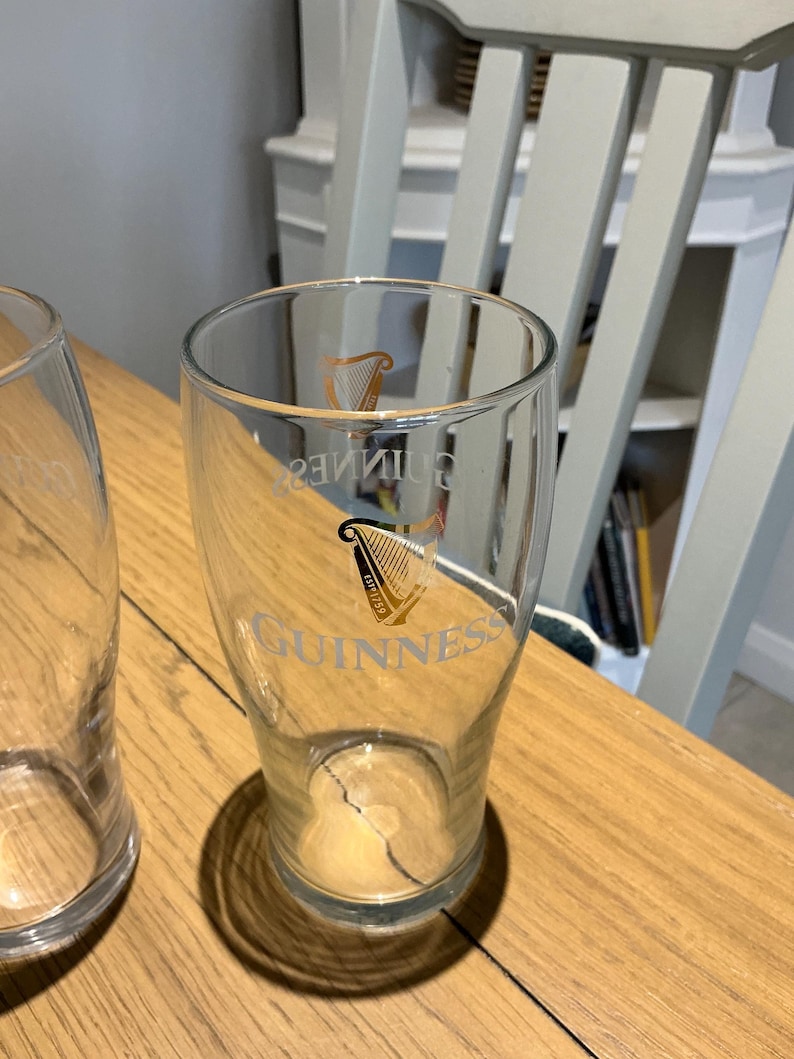 Puede incluir: Vaso de pinta transparente con el logotipo de Guinness, con un arpa dorada y la palabra "Guinness". El vaso est&aacute; sobre una superficie de madera, con un segundo vaso parcialmente visible.