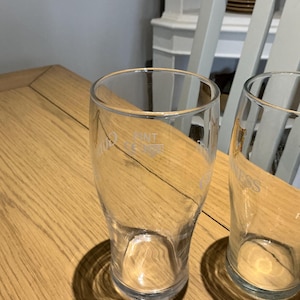 Puede incluir: Dos vasos de pinta transparentes sobre una mesa de madera. Los vasos tienen una forma ligeramente c&oacute;nica y est&aacute;n vac&iacute;os. Un vaso tiene la palabra "GUINNESS" impresa. El otro vaso tiene la palabra "NT".