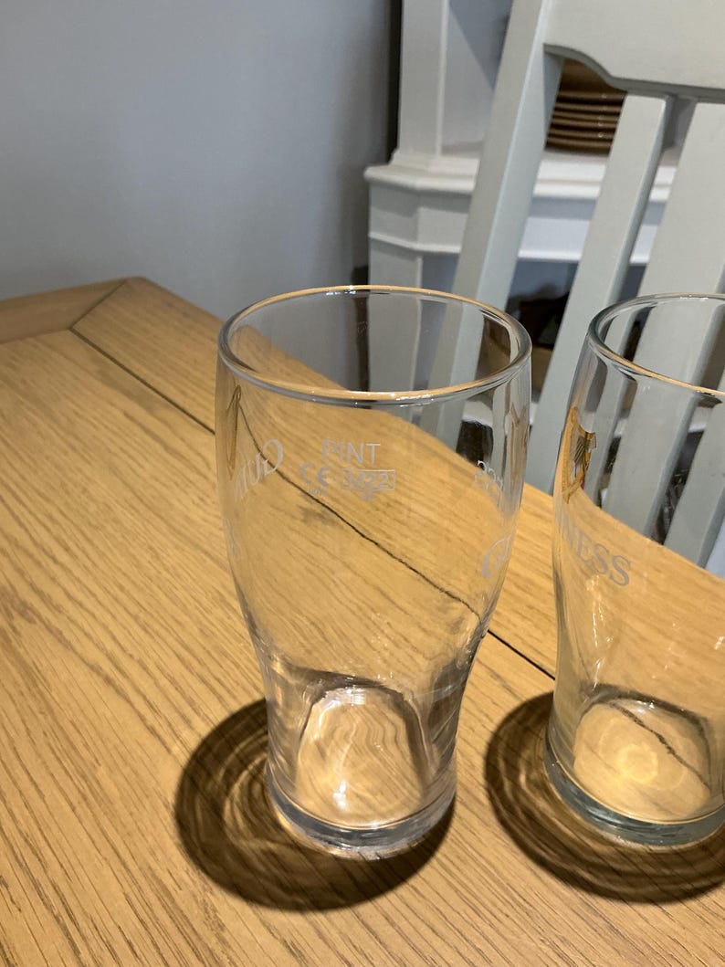 Puede incluir: Dos vasos de pinta transparentes sobre una mesa de madera. Los vasos tienen una forma ligeramente c&oacute;nica y est&aacute;n vac&iacute;os. Un vaso tiene la palabra "GUINNESS" impresa. El otro vaso tiene la palabra "NT".