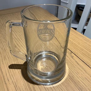 May include: A clear glass beer mug with a handle. The mug has a cylindrical body and a thick base. The Guinness logo is visible on the front of the mug. The mug is sitting on a wooden surface.