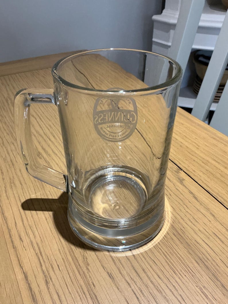 May include: A clear glass beer mug with a handle. The mug has a cylindrical body and a thick base. The Guinness logo is visible on the front of the mug. The mug is sitting on a wooden surface.
