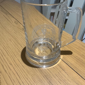 May include: A clear glass beer mug with a handle. The mug has a cylindrical shape and a thick base. The glass has etched text and a logo. The mug is sitting on a wooden surface.