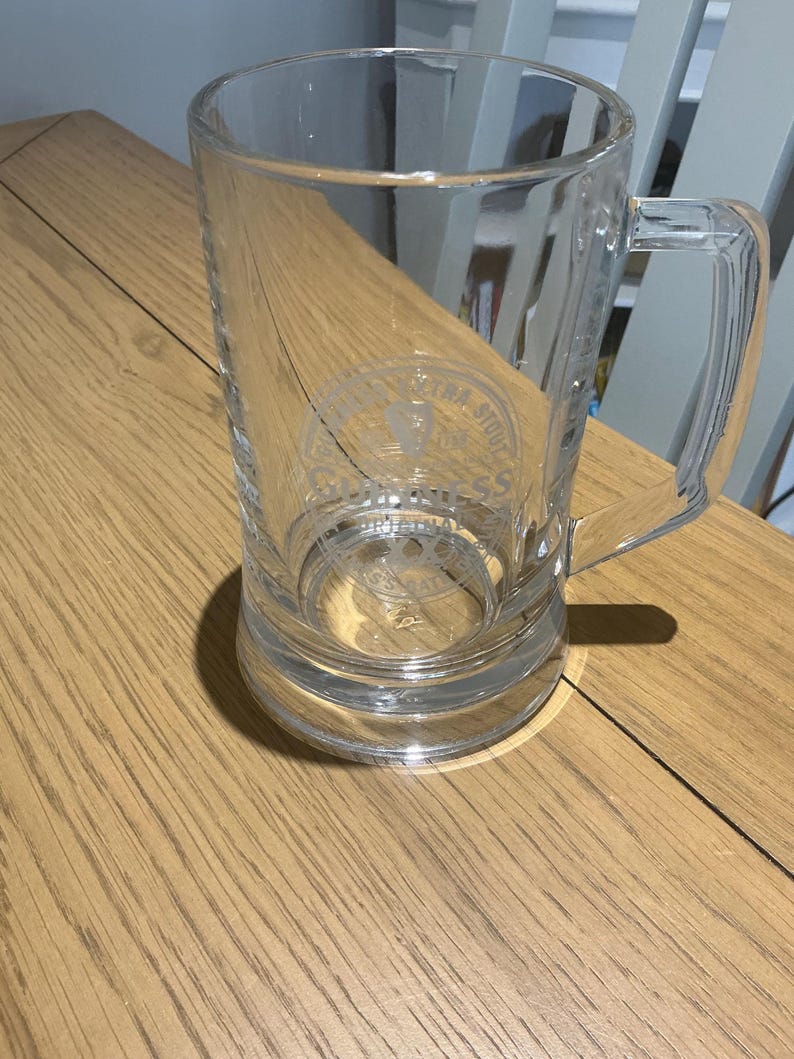May include: A clear glass beer mug with a handle. The mug has a cylindrical shape and a thick base. The glass has etched text and a logo. The mug is sitting on a wooden surface.