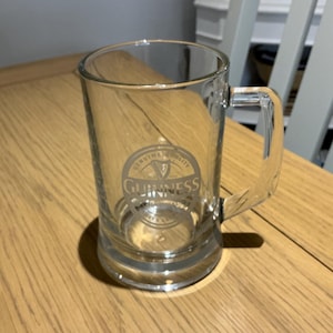 May include: A clear glass Guinness beer mug with a handle. The Guinness logo is printed on the front of the mug. The mug is sitting on a wooden table.
