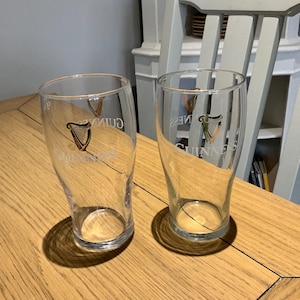 Puede incluir: Dos vasos de pinta transparentes con el logotipo de Guinness, con un arpa, est&aacute;n sobre una mesa de madera. Los vasos est&aacute;n vac&iacute;os y tienen forma curvada. El logotipo es dorado y negro.