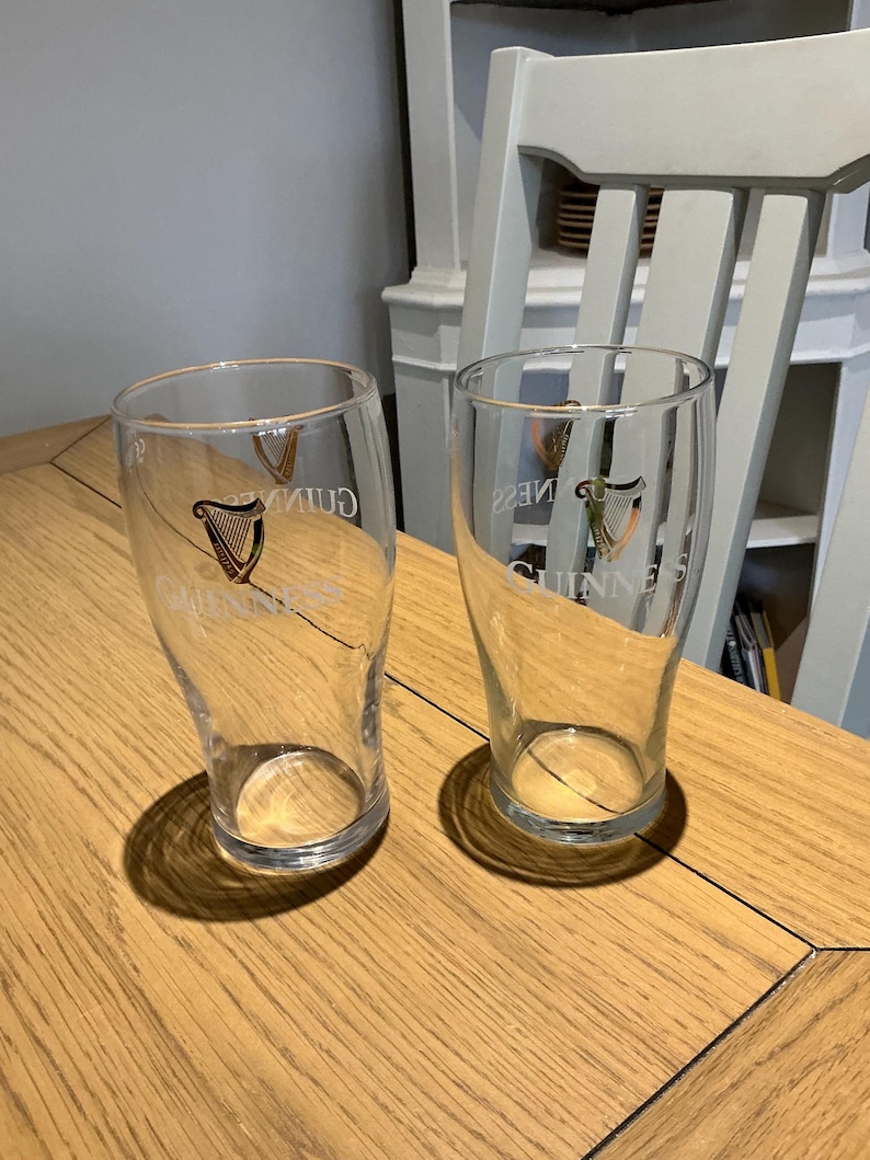 Puede incluir: Dos vasos de pinta transparentes con el logotipo de Guinness, con un arpa, est&aacute;n sobre una mesa de madera. Los vasos est&aacute;n vac&iacute;os y tienen forma curvada. El logotipo es dorado y negro.