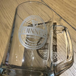 May include: A clear glass beer mug with a rectangular shape and a handle. The Guinness logo is etched on the front. The logo includes the word "Guinness" in a circular design with the words "Original" and "James's Gate Dublin".