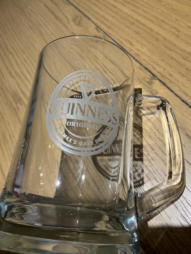 May include: A clear glass beer mug with a rectangular shape and a handle. The Guinness logo is etched on the front. The logo includes the word "Guinness" in a circular design with the words "Original" and "James's Gate Dublin".