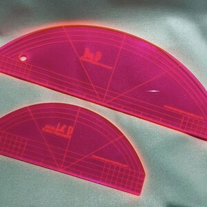 May include: Two neon pink acrylic quilting templates with curved edges and grid markings. The larger template has the text "Jay D Curves & More" and the smaller one has "Jay D Curves & More". Both have a hole for hanging.