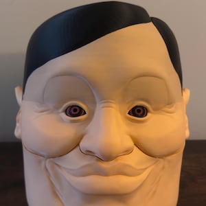 May include: A large, smiling face sculpture with a black hairpiece. The face is a light beige color with dark red eyes. The sculpture has a wide, exaggerated smile and a smooth, rounded shape. The overall design is cartoonish and somewhat unsettling.