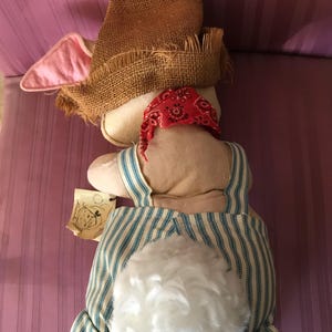 May include: A stuffed bunny doll wearing a burlap hat, red bandana, and blue and white striped overalls. The bunny has a fluffy white tail and pink-lined ears. A small tag with a drawing is attached.