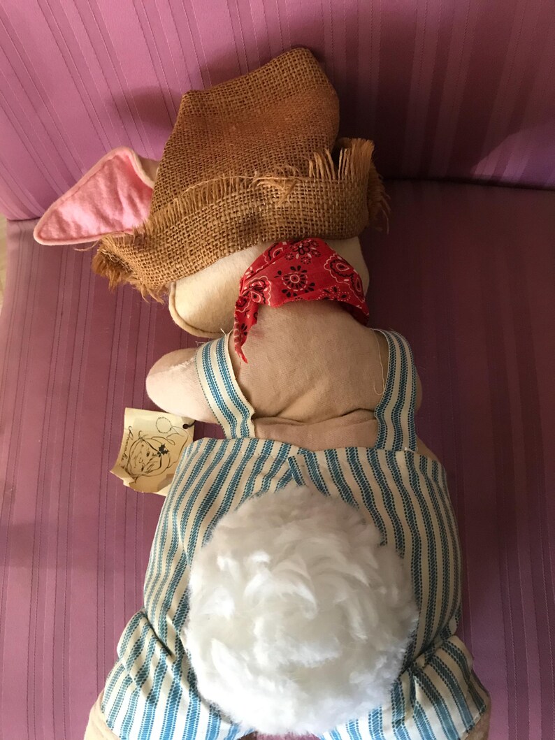May include: A stuffed bunny doll wearing a burlap hat, red bandana, and blue and white striped overalls. The bunny has a fluffy white tail and pink-lined ears. A small tag with a drawing is attached.