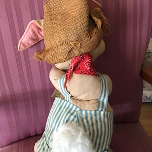 May include: A stuffed bunny wearing a burlap hat, red bandana, and blue and white striped overalls. The bunny has pink ears and a fluffy white tail. The bunny is sitting on a pink striped chair.
