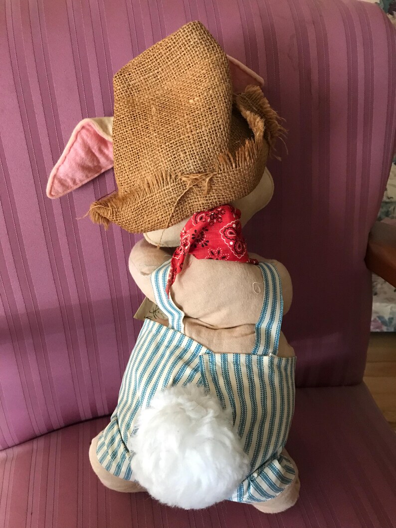 May include: A stuffed bunny wearing a burlap hat, red bandana, and blue and white striped overalls. The bunny has pink ears and a fluffy white tail. The bunny is sitting on a pink striped chair.