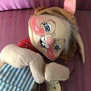 May include: A handmade, whimsical rabbit doll with a painted face, blue eyes, and a pink nose. The doll wears a burlap hat, a red bandana, and blue and white striped overalls. The doll has long, floppy ears and a tag.
