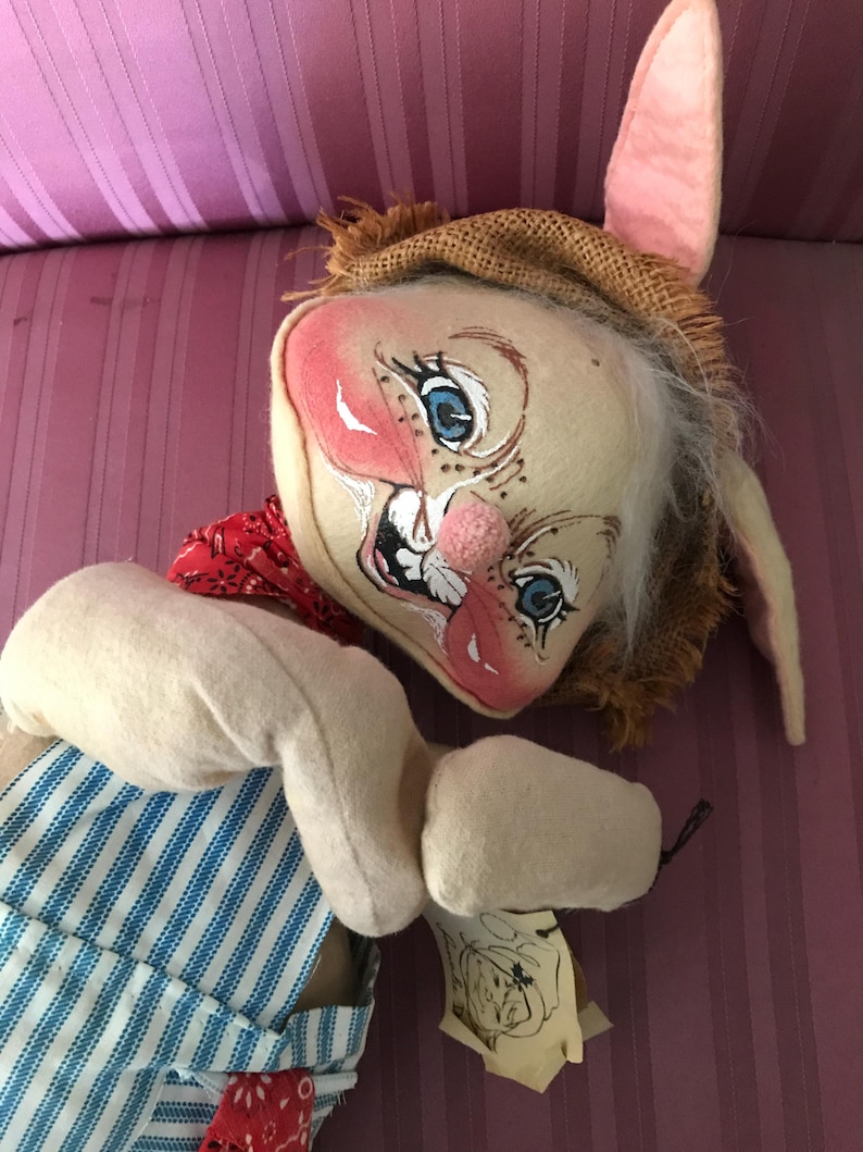 May include: A handmade, whimsical rabbit doll with a painted face, blue eyes, and a pink nose. The doll wears a burlap hat, a red bandana, and blue and white striped overalls. The doll has long, floppy ears and a tag.