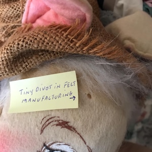 May include: A handmade plush bunny doll with a burlap hat and pink felt ears. The doll has painted facial features, including blue eyes and rosy cheeks. A yellow sticky note reads "Tiny divot in felt manufacturing."