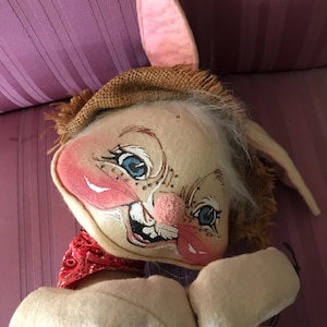 May include: A whimsical, handmade bunny doll with a painted face, blue eyes, and a pink nose. The bunny wears a straw hat, a red bandana, and a blue and white striped outfit. The doll has long, floppy ears and white fur.