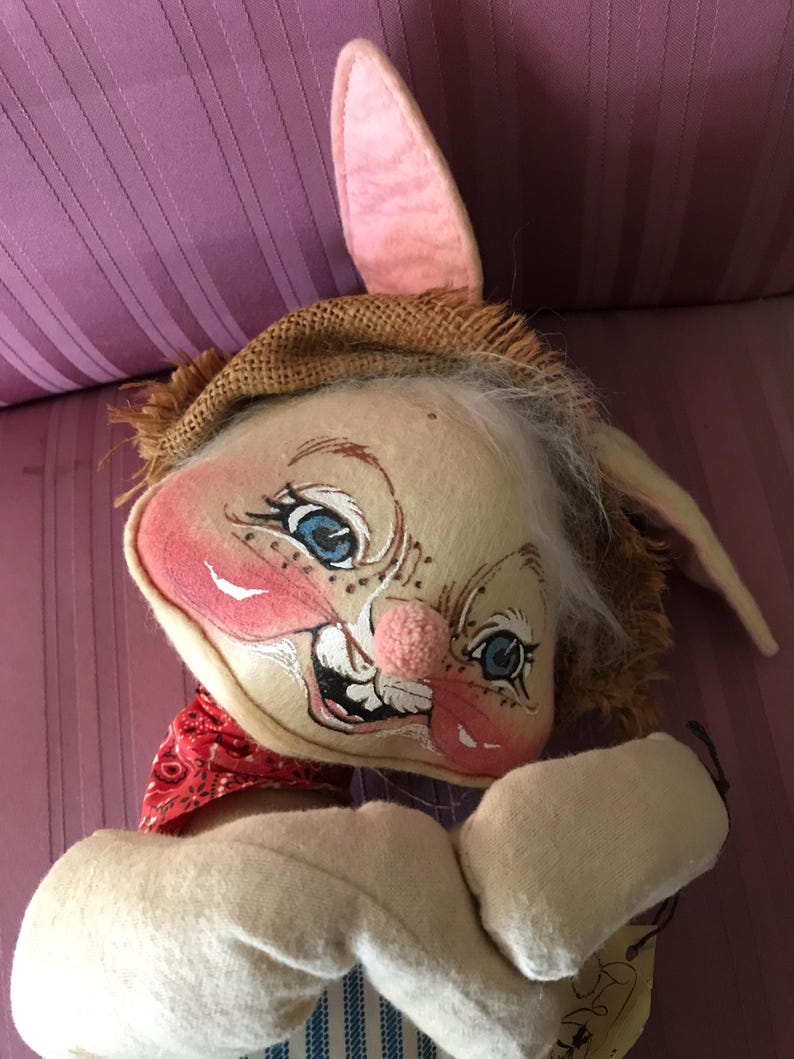 May include: A whimsical, handmade bunny doll with a painted face, blue eyes, and a pink nose. The bunny wears a straw hat, a red bandana, and a blue and white striped outfit. The doll has long, floppy ears and white fur.