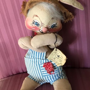 May include: A vintage stuffed bunny doll with a burlap hat and blue and white striped overalls. The bunny has a painted face with blue eyes, rosy cheeks, and a red neckerchief. A tag is attached to the doll.