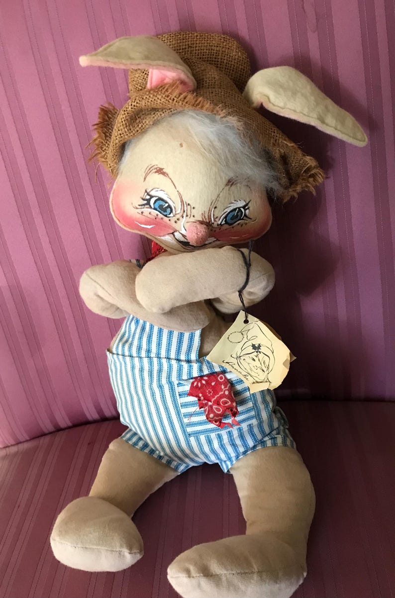 May include: A vintage stuffed bunny doll with a burlap hat and blue and white striped overalls. The bunny has a painted face with blue eyes, rosy cheeks, and a red neckerchief. A tag is attached to the doll.