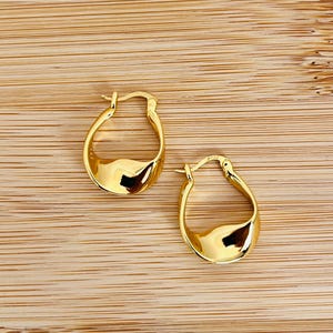 Peut inclure: Une paire de boucles d'oreilles créoles dorées. Les boucles d'oreilles présentent un design incurvé unique avec une surface lisse et réfléchissante. Elles sont présentées sur une surface en bois brun clair.