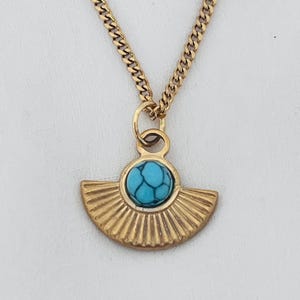 Può includere: Una collana color oro con un ciondolo a forma di ventaglio. Il ciondolo presenta una pietra rotonda di colore turchese al centro. La catena è a maglia curb. Lo sfondo è un tessuto bianco liscio.