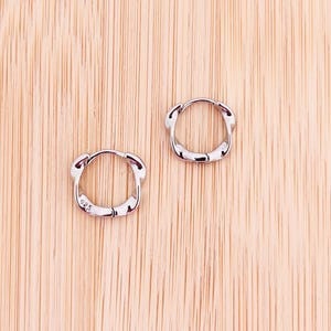 Puede incluir: Un par de pequeños aros de plata pulida con un diseño liso y redondeado. Cada pendiente cuenta con un mecanismo de cierre de bisagra y está claramente marcado con el número 925 en la superficie interior, indicando plata de ley.