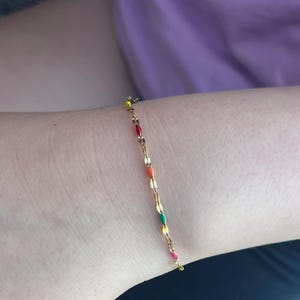 Könnte beinhalten: Ein zartes goldenes Armband mit einer Reihe von farbenfrohen, emaillierten ovalen Perlen. Die Perlen sind in den Farben Rot, Orange, Grün und Pink gehalten und verleihen dem Schmuck eine verspielte Note. Das Armband wird am Handgelenk getragen.