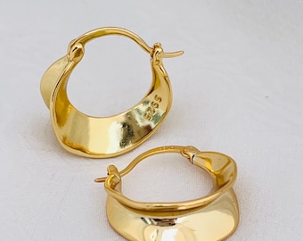 May include: A pair of gold-colored hoop earrings. The earrings have a unique, curved design with a smooth, reflective surface. The earrings are displayed on a white background, highlighting their polished finish. The earrings are made of a shiny, gold-toned metal.