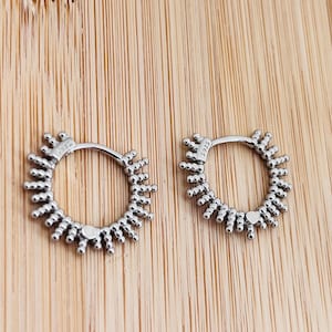 May include: A pair of silver hoop earrings featuring a sunburst design with small metallic beads radiating from the outer edge. Each earring is stamped with the text 925, indicating sterling silver construction, and has a hinged closure mechanism.
